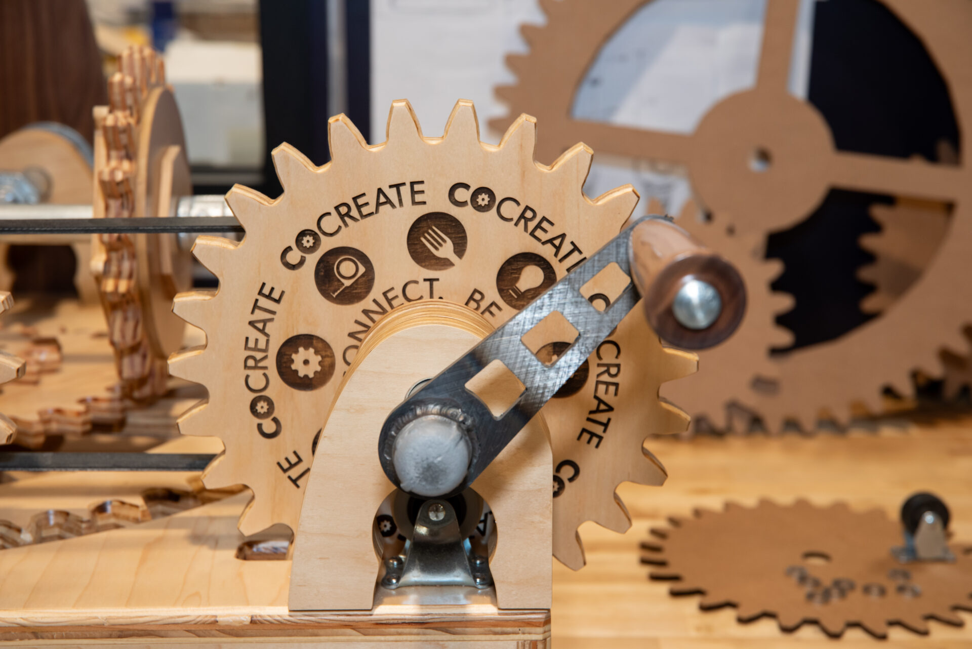 Wooden gear with metal hand crank with the CoCreate logo on it.